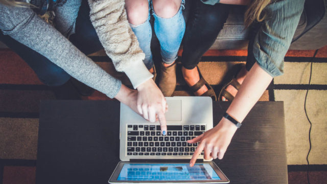 Three women sitting on a couch pointing at a laptop screen, overhead shot shows legs and arms only - photo credit @johnschno